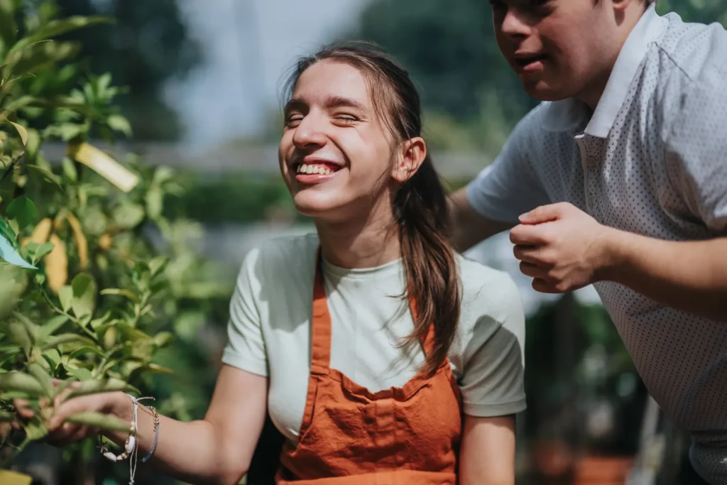 Woman in wheelchair in an outdoor garden smiling while soaking in the sunlight. She is accompanied by a young man with down syndrome
