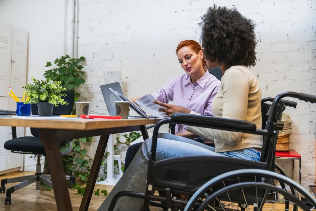 Woman in a wheelchair and her female colleague looking over paperwork in an office setting