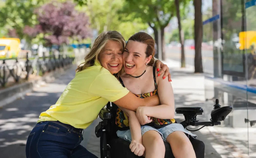 Young woman in a wheelchair and her female support worker who is crouching down to give her a hug both smiling and laughing at the camera